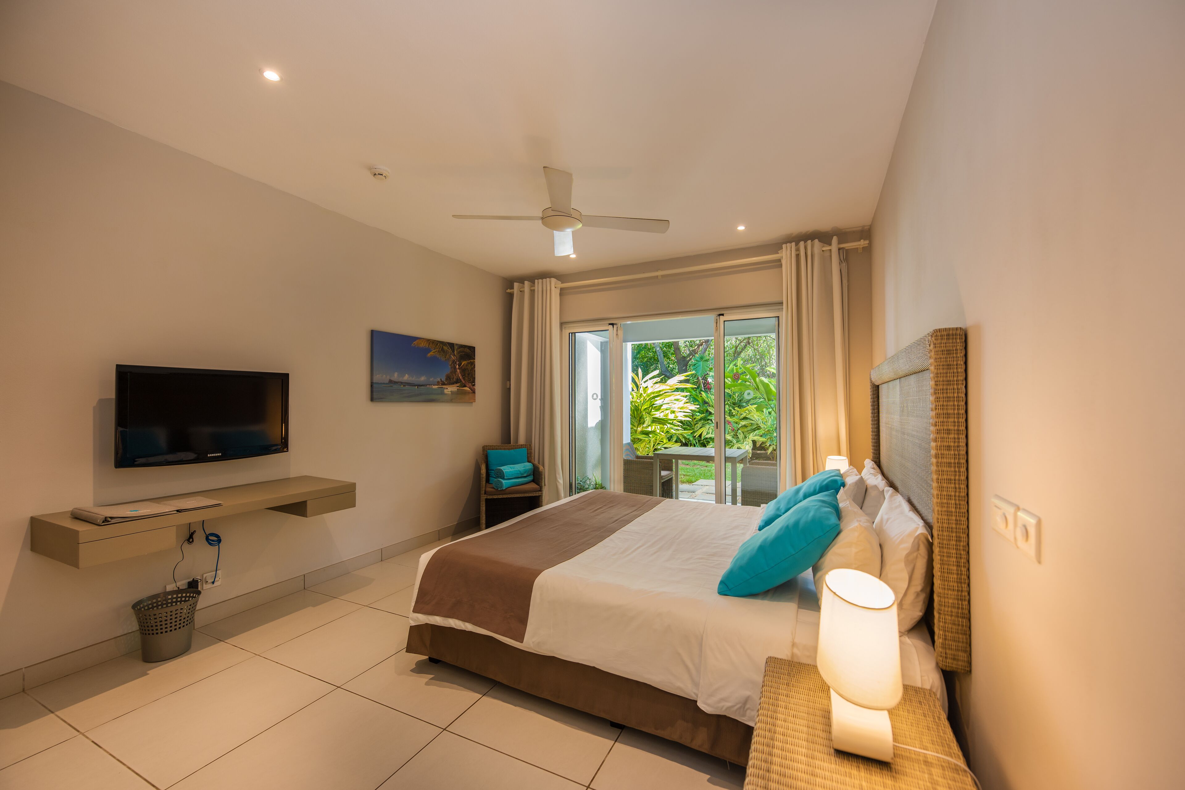 Habitación de hotel con cama, televisor, ventilador de techo, ventana con cortinas y vistas al exterior.