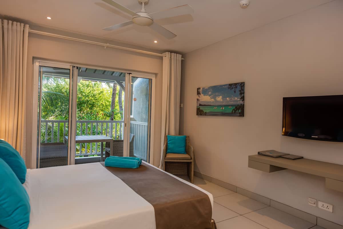 Habitación de hotel con cama, una silla, televisor y balcón con vistas a zonas verdes.