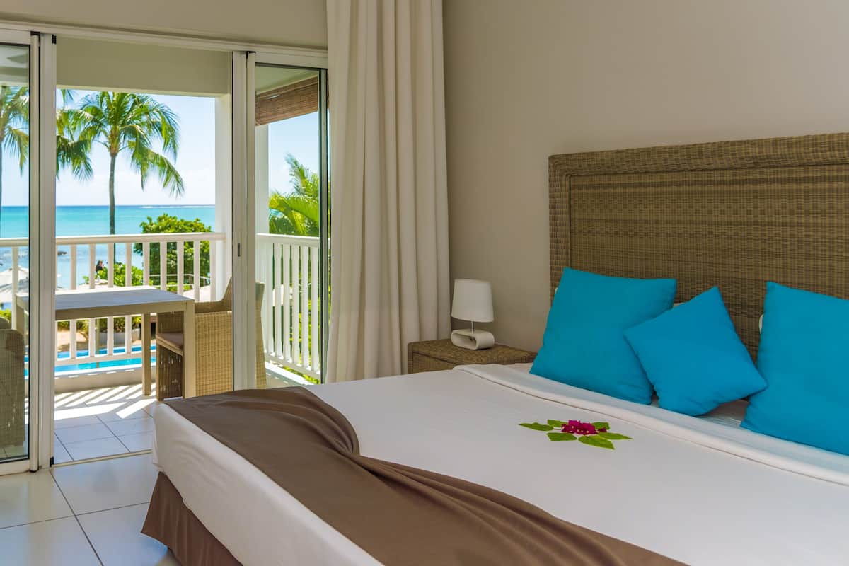 Habitación de hotel con cama, mesita de noche, lámpara y vista al océano y palmeras a través de las puertas del balcón.
