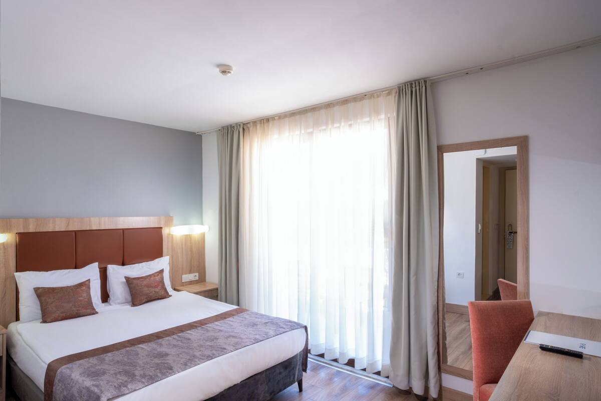 A hotel room with a bed, a desk, a chair, a mirror, and a window with curtains.