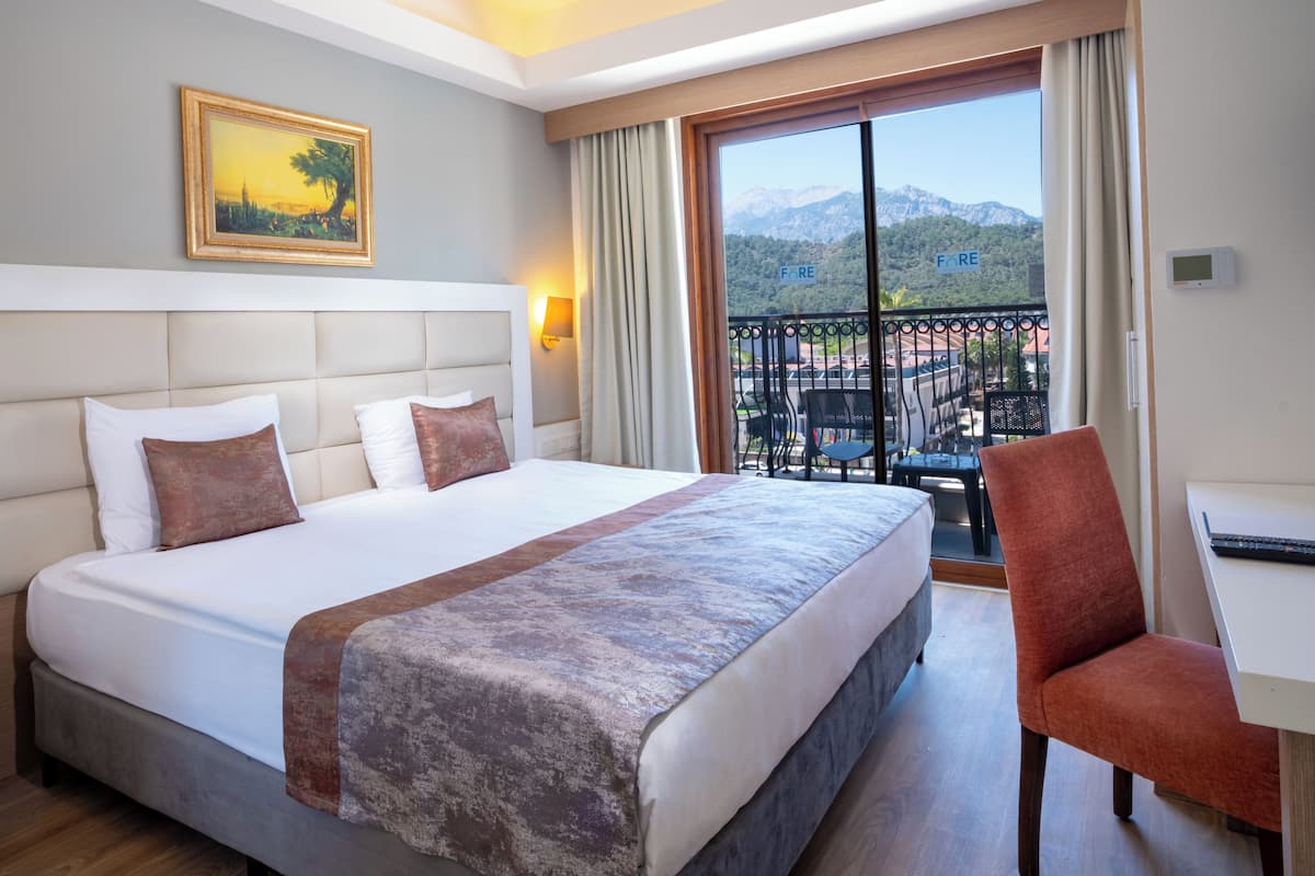 A hotel room with a large bed, a desk, and a balcony with a view of mountains.