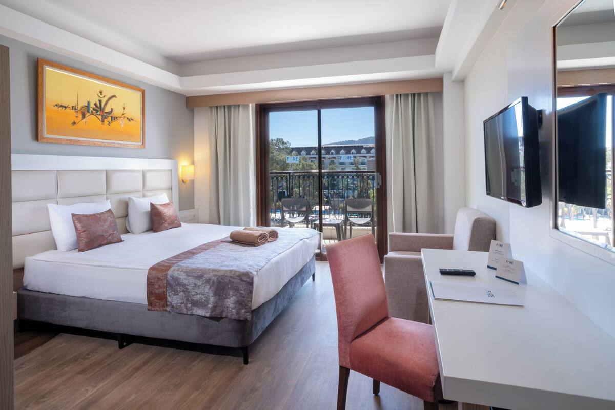 A hotel room with a bed, a desk, a chair, a TV, and a balcony with a view of buildings.
