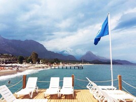 Ένα κατάστρωμα με λευκές ξαπλώστρες που προσφέρει θέα σε μια παραλία και μια προβλήτα, με βουνά στο βάθος.