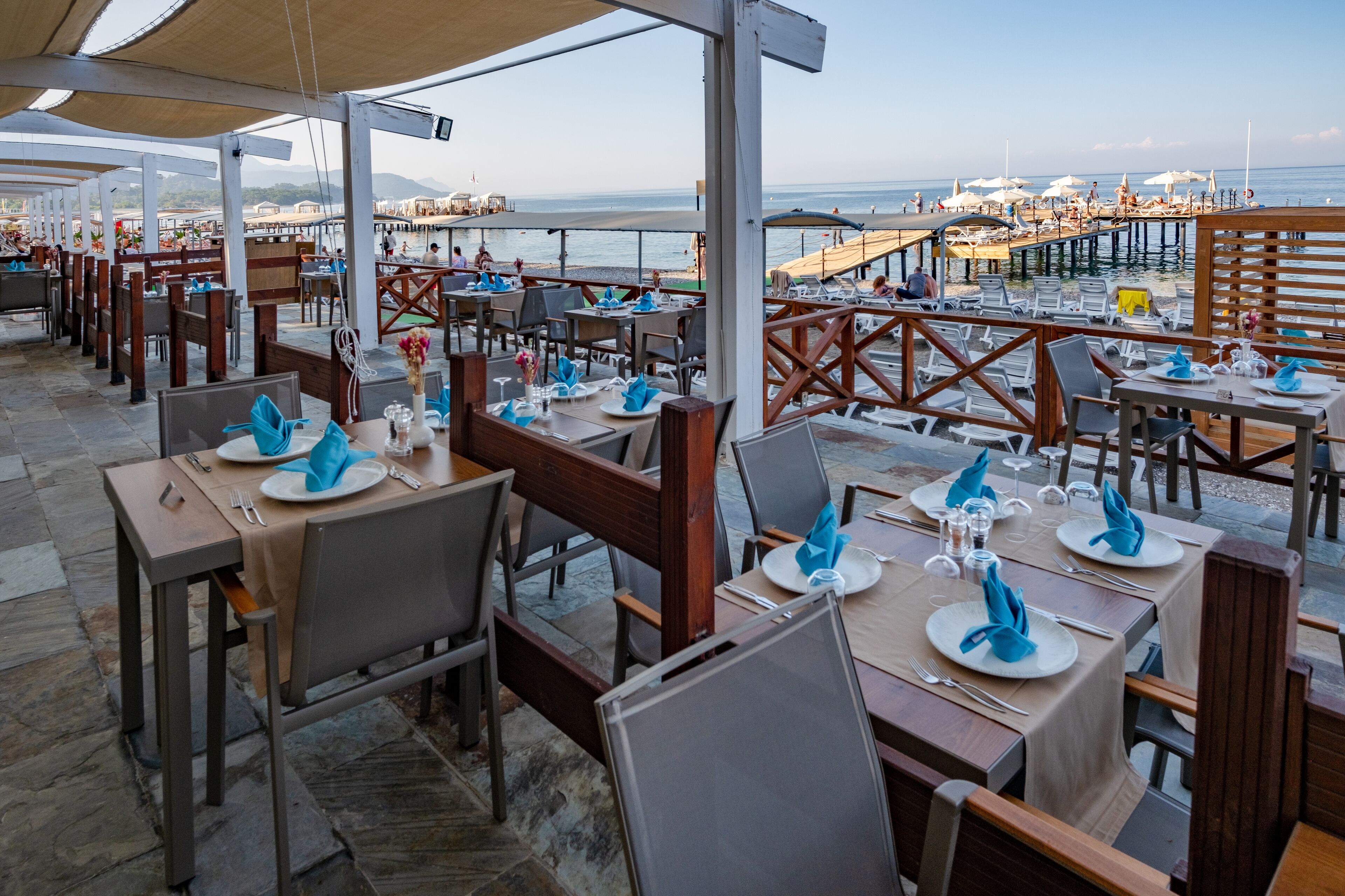 Un área de comedor al aire libre con mesas dispuestas para dos, con vista a una playa y un muelle.