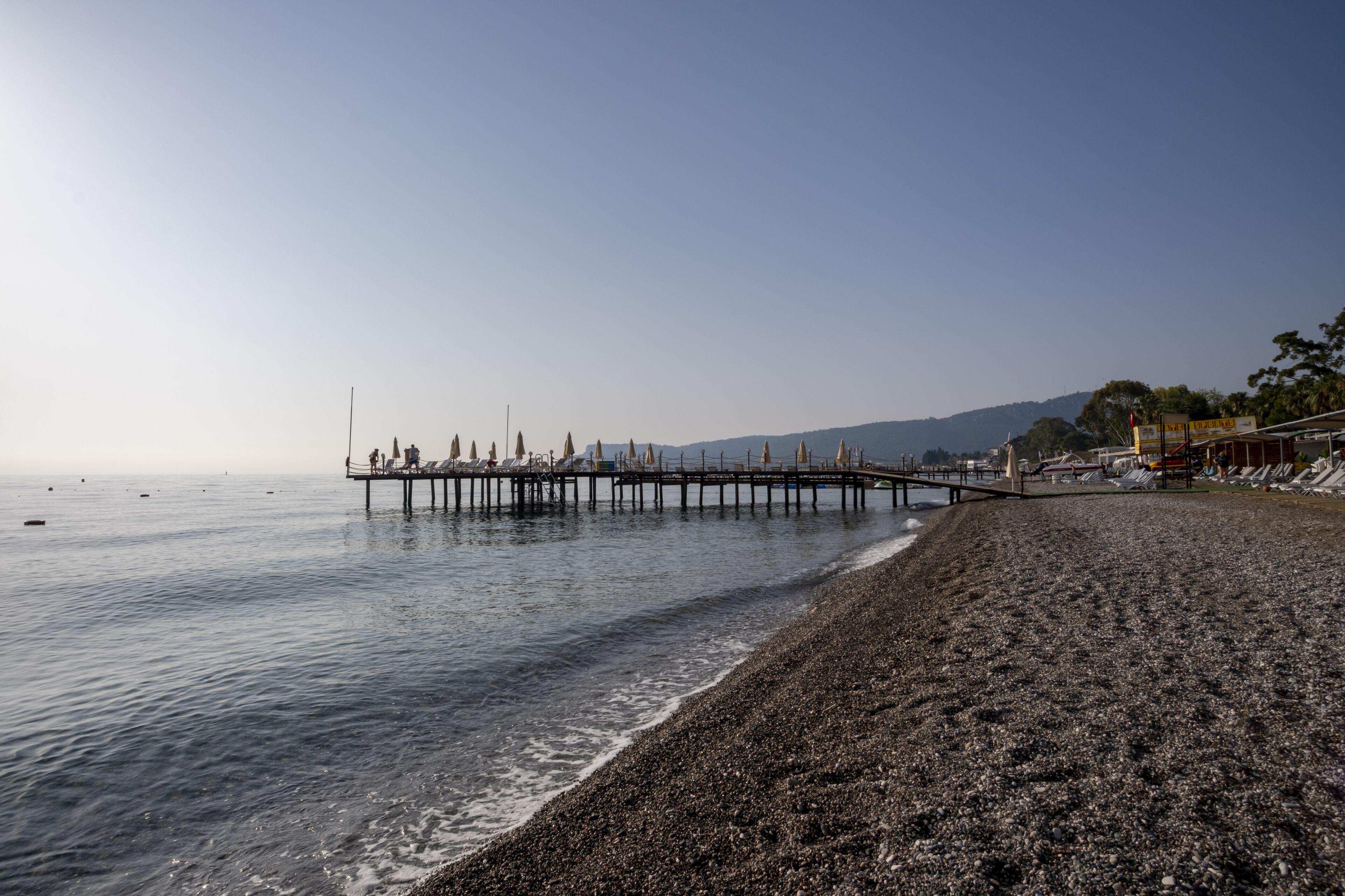 A pebbled beach with a pier extending into calm waters, surrounded by hills.
