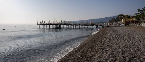 A pebbled beach with a pier extending into calm waters, surrounded by hills.
