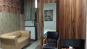 A room with a beige sofa, a black chair, a wooden wardrobe, and a painting on the wall. - Hotel RTS Plaza Near IGI Airport (New Delhi)