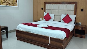A neatly made bed with a wooden frame, white bedding, and red pillows. There is a small wooden nightstand with a phone on it to the right of the bed. - Hotel RTS Plaza Near IGI Airport (New Delhi)