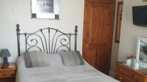 A neatly made bed with a metal headboard, a wooden door, a nightstand with a lamp, and a dresser with a mirror. - Bryn Coed Guest House (Colwyn Bay)