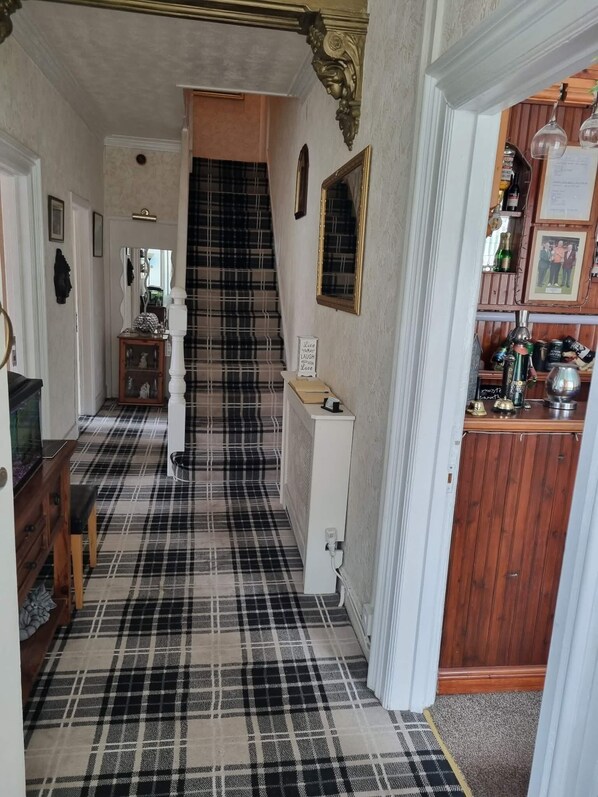 A hallway with a checkered floor, a staircase, a mirror, a sign, and a bar area with bottles and glasses. - Bryn Coed Guest House (Colwyn Bay)