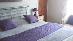 A bed with a headboard, two pillows, a purple blanket, a nightstand with a lamp, and a wooden door. - Bryn Coed Guest House (Colwyn Bay)