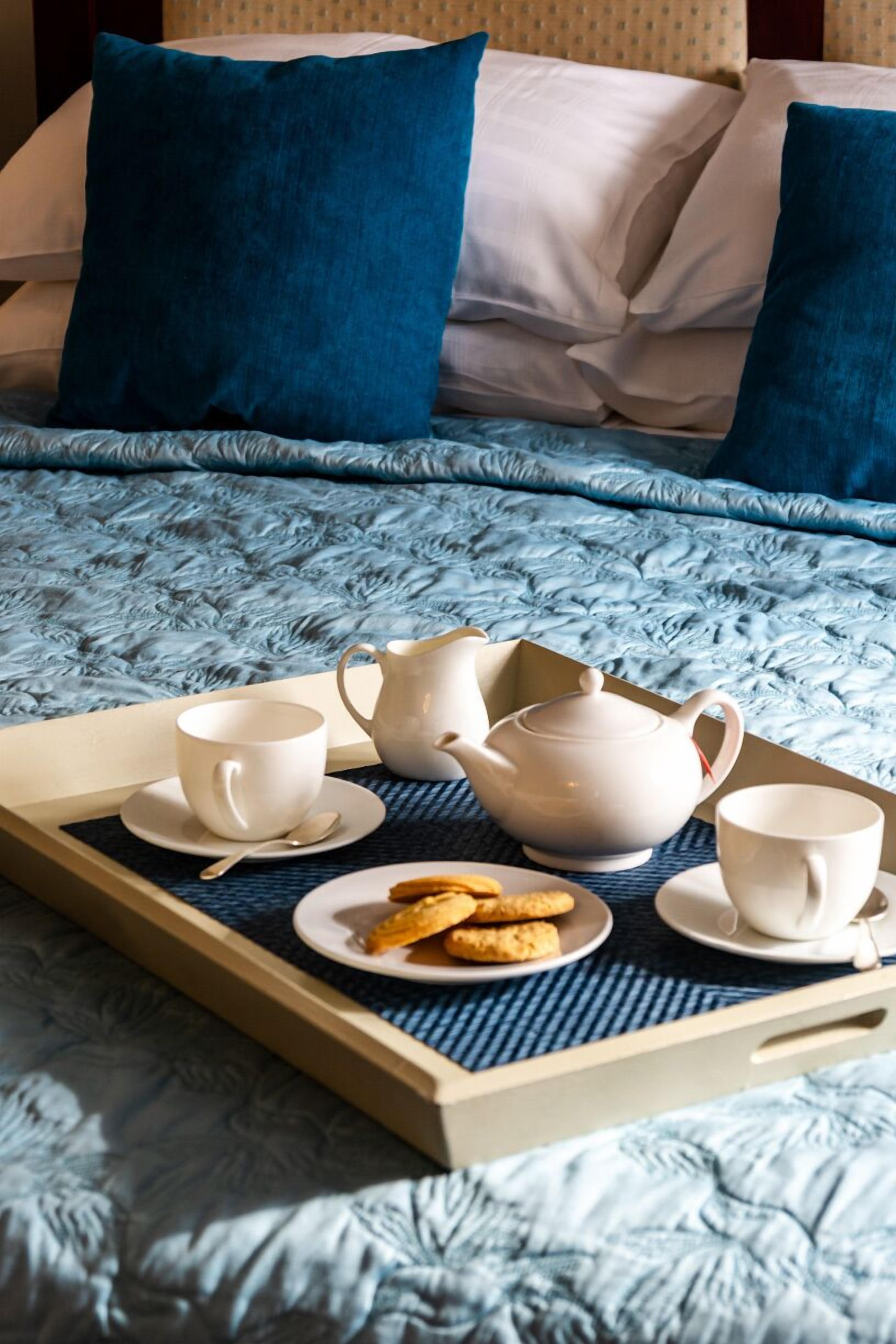 Una cama bien hecha con colcha azul, dos almohadas, una bandeja con juego de té y galletas, y una taza blanca.