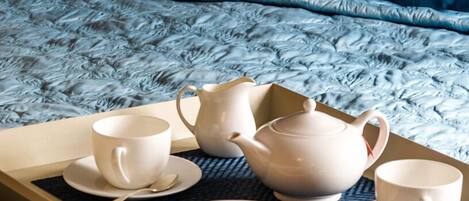 Una cama bien hecha con colcha azul, dos almohadas, una bandeja con juego de té y galletas, y una taza blanca.