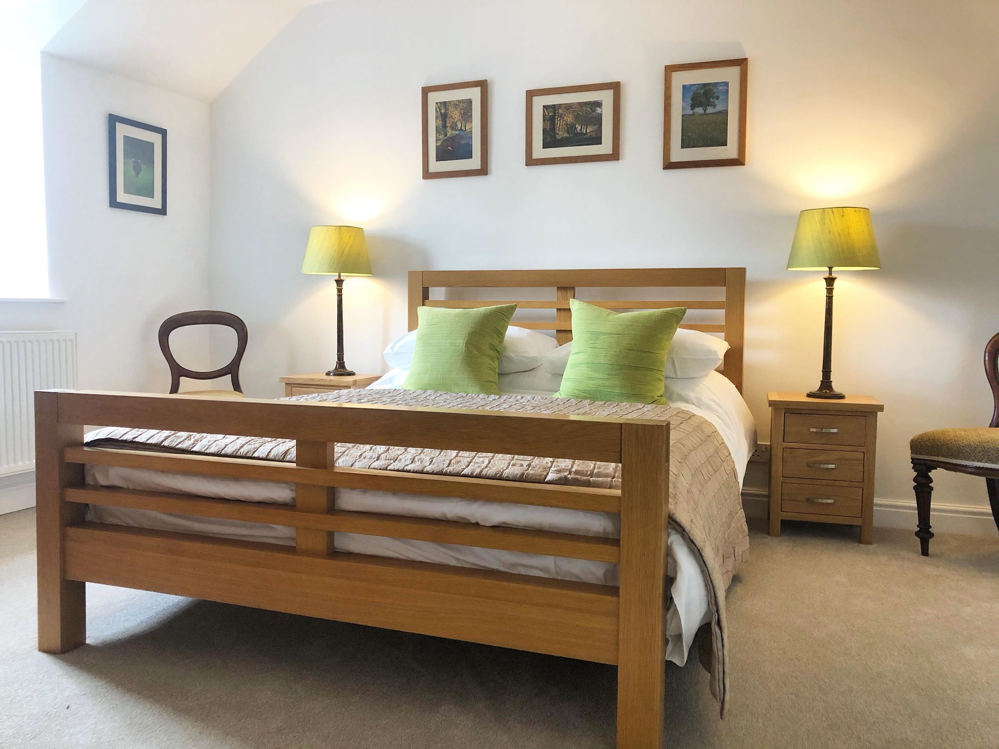 A bedroom with a wooden bed, two bedside tables with lamps, a chair, and three framed pictures on the wall.