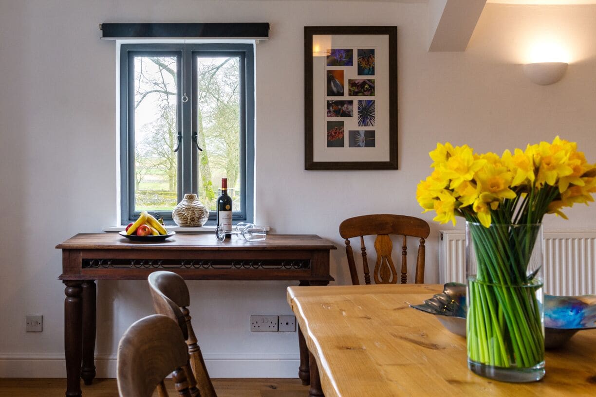A dining area with a wooden table, chairs, a vase of yellow flowers, a window with a view of trees, and a framed collage on the wall.