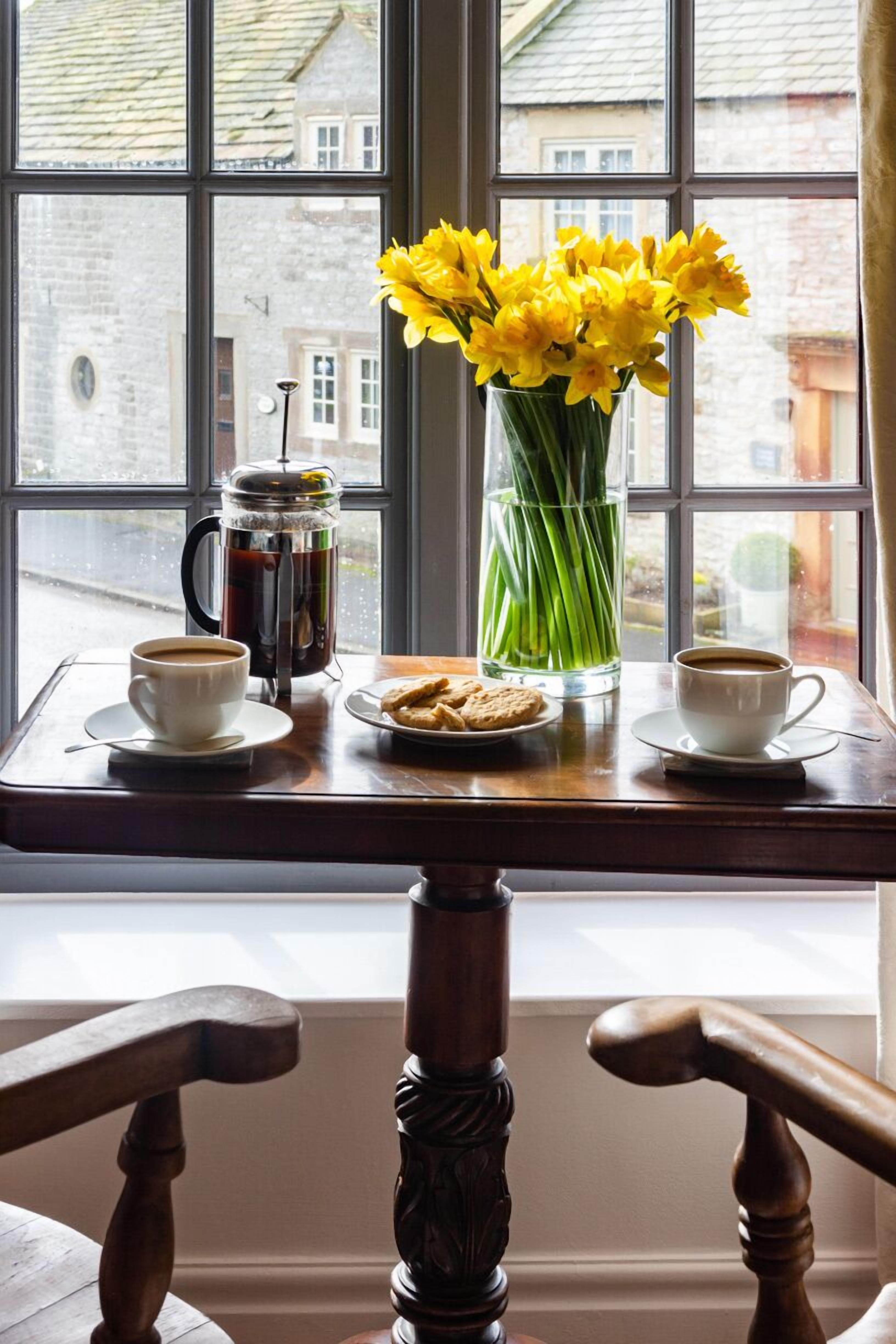 Un tavolo con una caffettiera, due tazze di caffè, un piatto di biscotti e un vaso di fiori gialli vicino a una finestra.