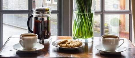 A table with a coffee pot, two cups of coffee, a plate of cookies, and a vase of yellow flowers by a window.