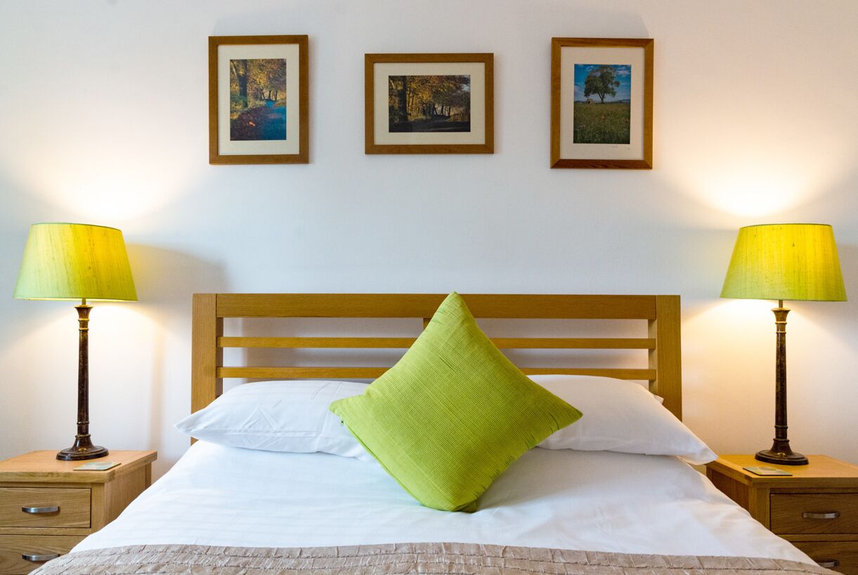 Una cama bien hecha con un almohadón verde, dos lámparas de noche y tres cuadros en la pared.