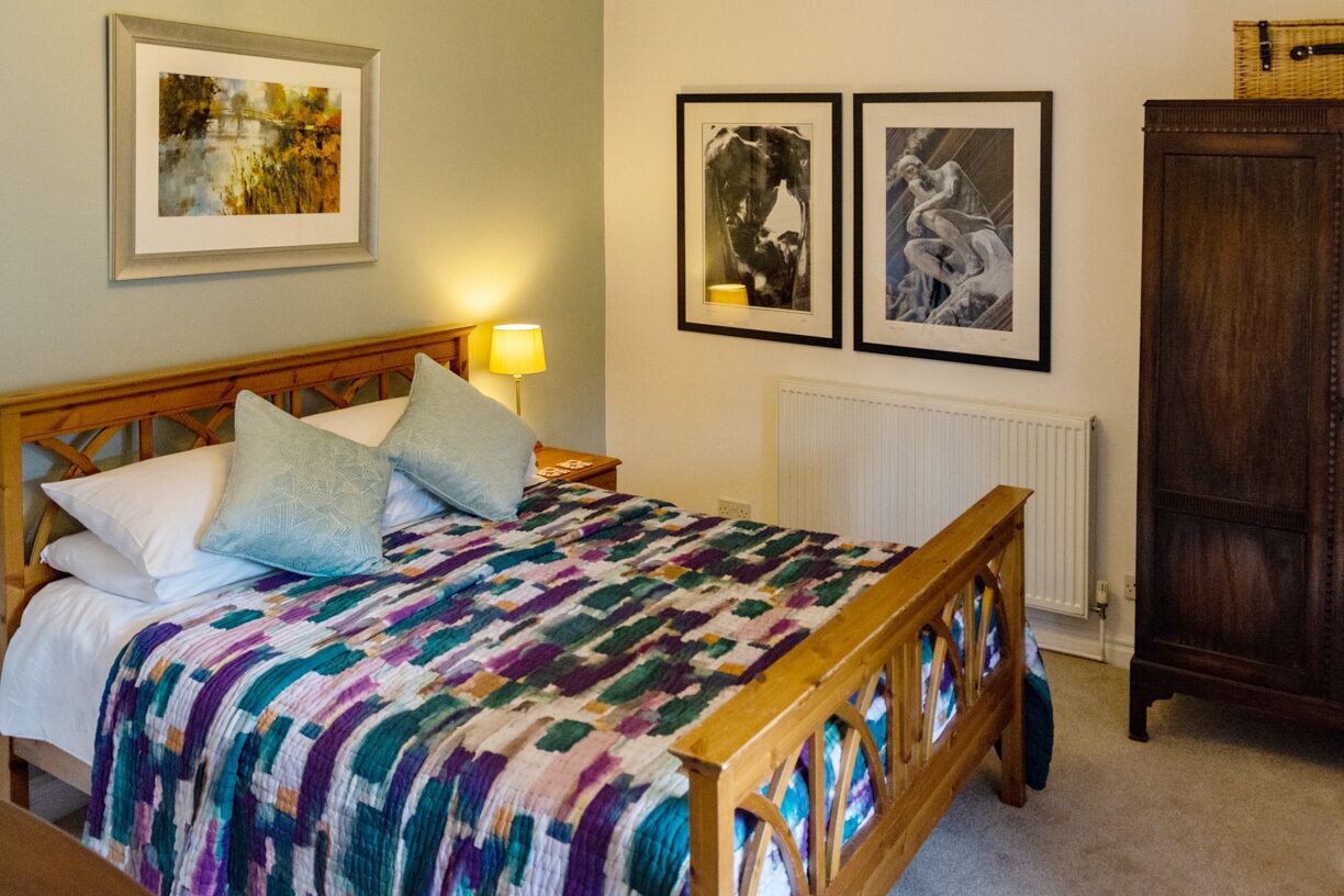 A bed with a patterned blanket, a wooden headboard, and two pillows. There are framed pictures on the wall, a lamp on the nightstand, and a wooden cabinet.