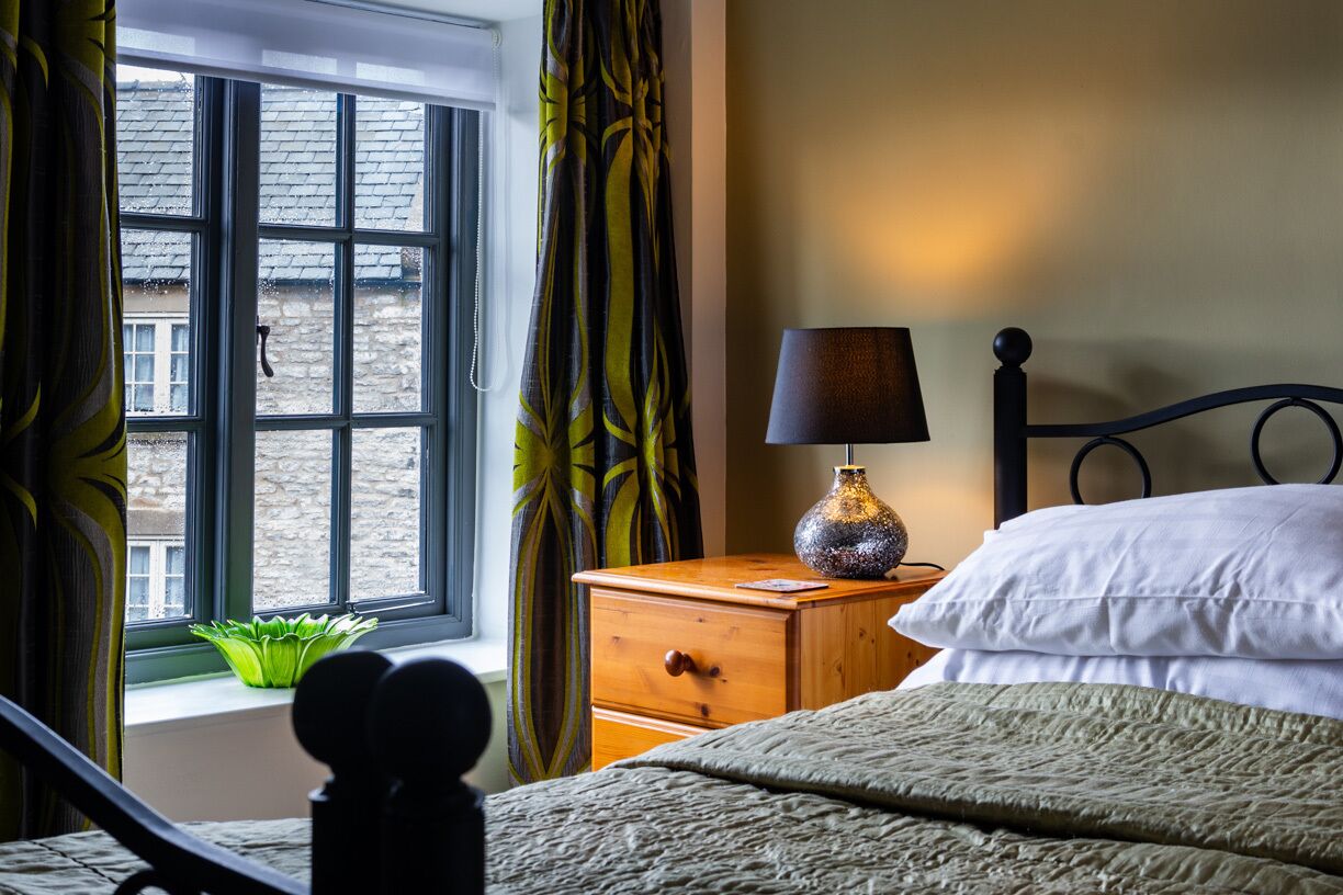 A bedroom with a bed, a nightstand with a lamp, a window with curtains, and a view of the outside.