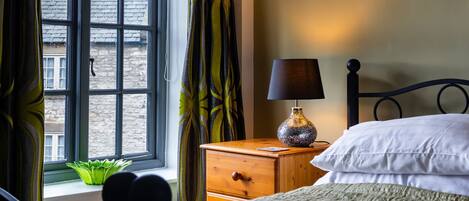 A bedroom with a bed, a nightstand with a lamp, a window with curtains, and a view of the outside.