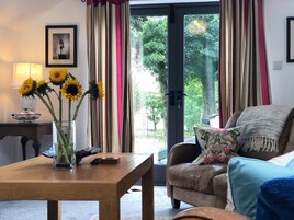 Una sala de estar con una mesa de madera, un jarrón con girasoles, un sofá con un cojín floral y vistas a zonas verdes a través de un ventanal.