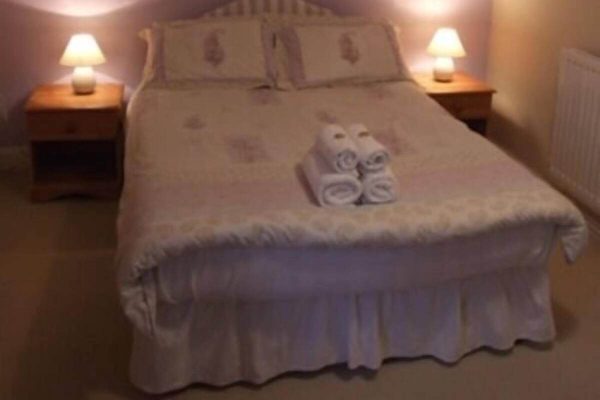 A neatly made bed with a white duvet, two pillows, and rolled towels on the bed. There are two bedside tables with lamps on either side of the bed.