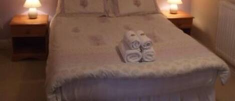 Una cama bien hecha con edredón blanco, dos almohadas y toallas enrolladas. Hay dos mesitas de noche con lámparas a cada lado de la cama.