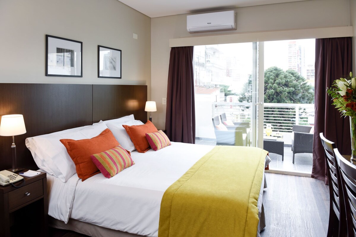 A hotel room with a bed, two bedside lamps, a window with curtains, and a view of the outside.