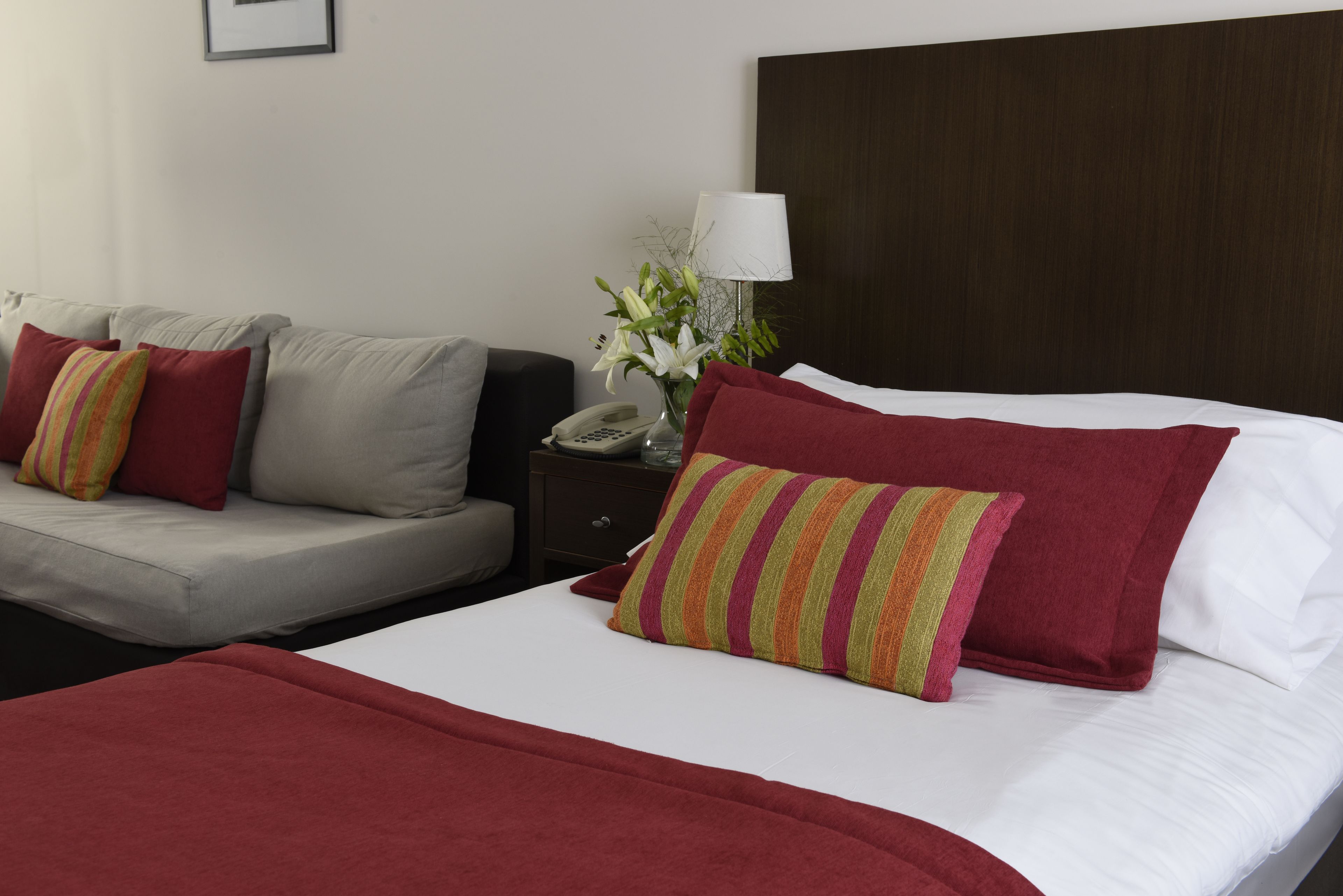 A hotel room with a bed, a sofa, a nightstand with a lamp, and a vase with flowers.