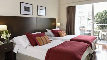 A hotel room with two beds, a nightstand, a vase with flowers, and a balcony with a view of the city.