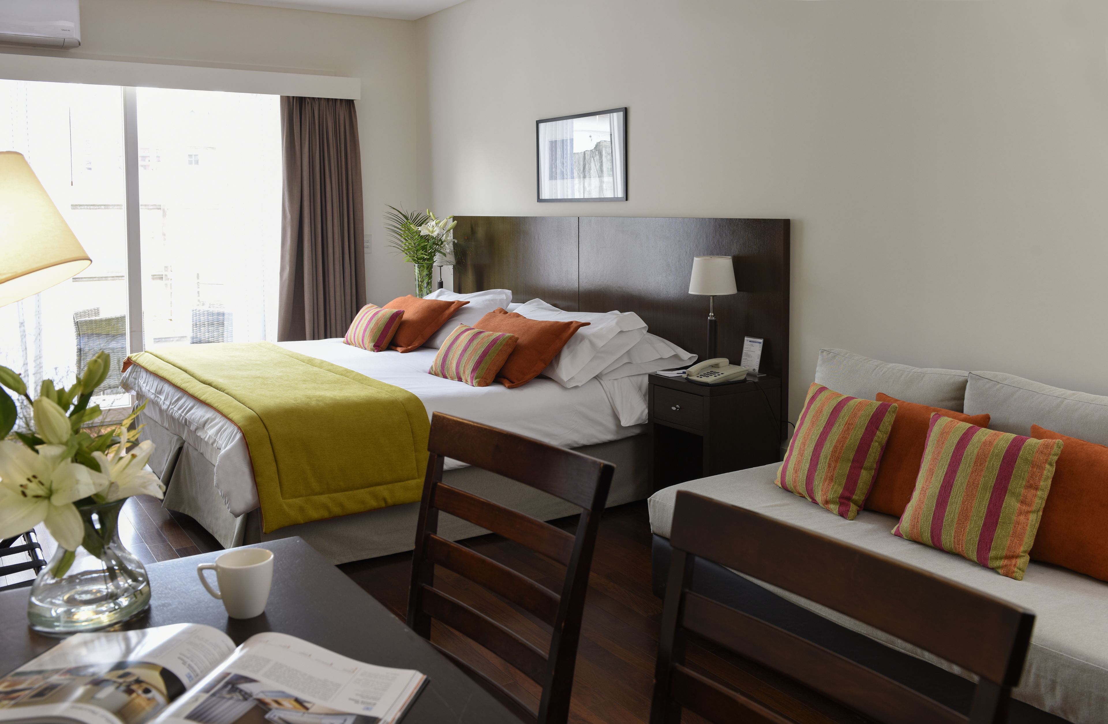 Une chambre d’hôtel avec un lit, un canapé, un bureau avec une lampe, un vase de fleurs et une fenêtre avec des rideaux.