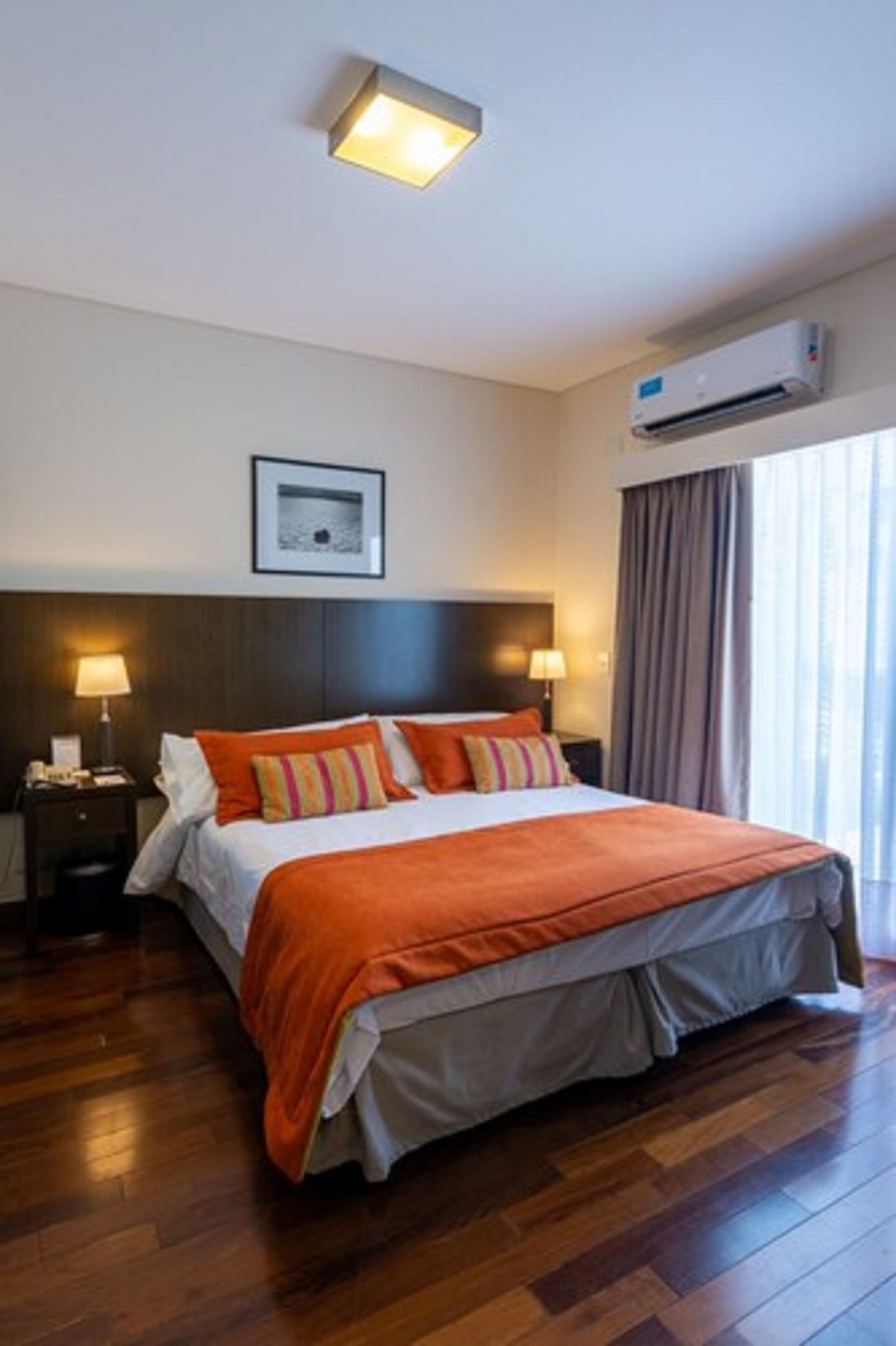 A hotel room with a large bed, a nightstand, a wall-mounted air conditioner, and a window with curtains.