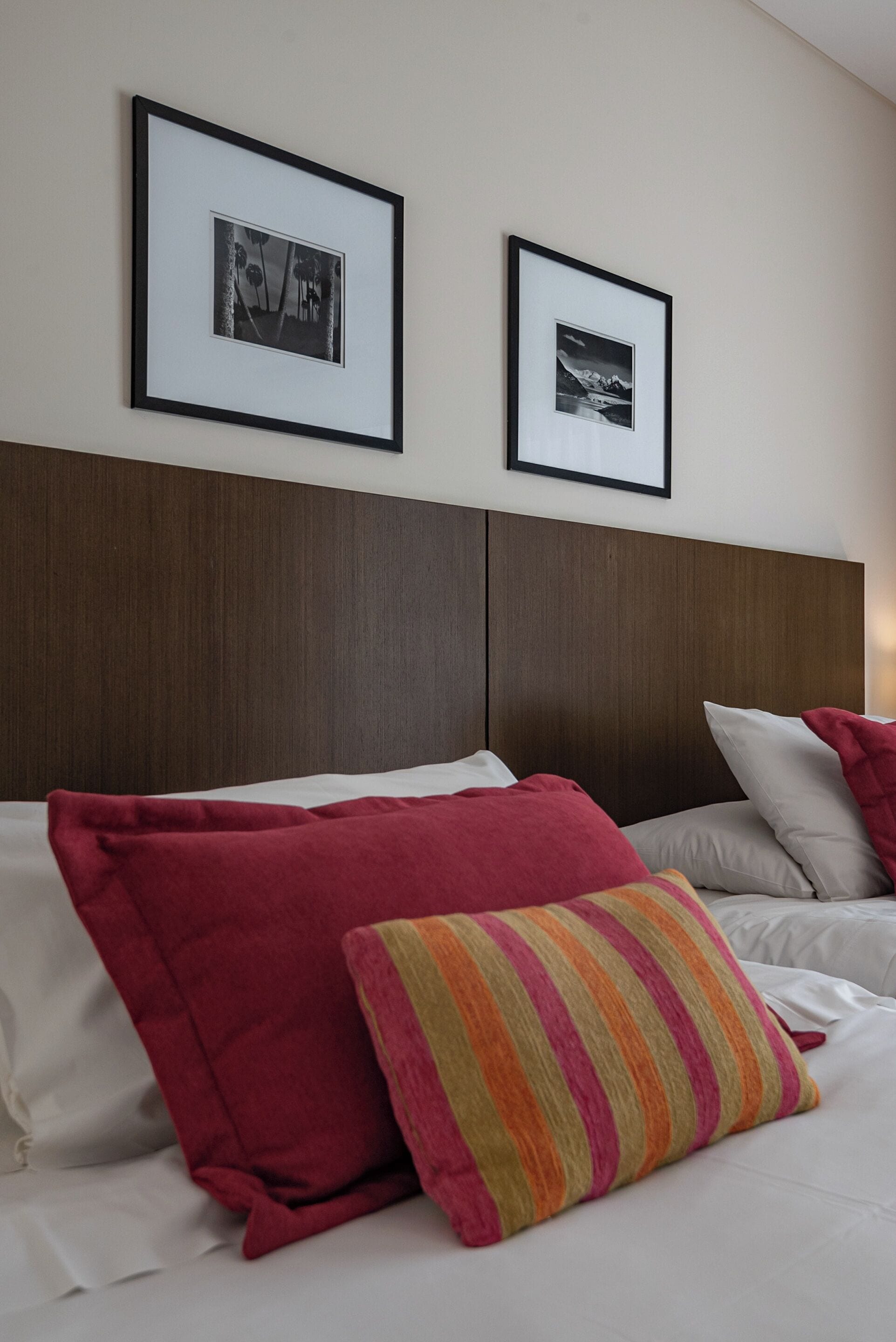 Una camera d'albergo con una testiera in legno, due quadri appesi al muro e un letto con lenzuola bianche e cuscini colorati.