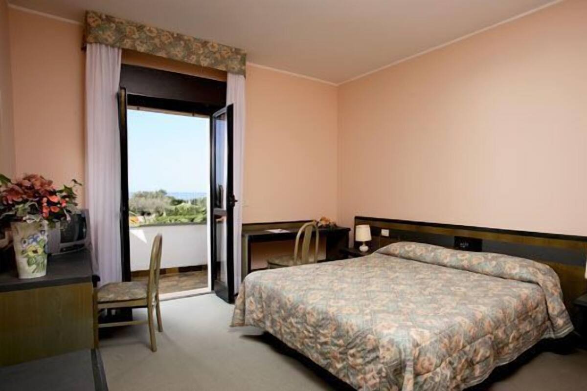 A hotel room with a bed, a desk, a chair, a lamp, and a view of the outdoors.