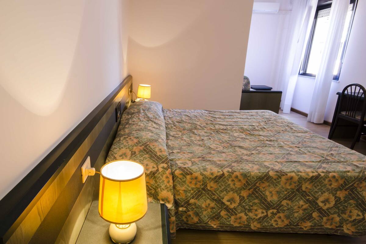 A hotel room with a bed, a bedside lamp, a chair, and a window with curtains.