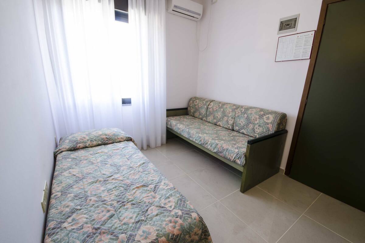 A room with two beds, a sofa, a window with curtains, an air conditioner, and a door.
