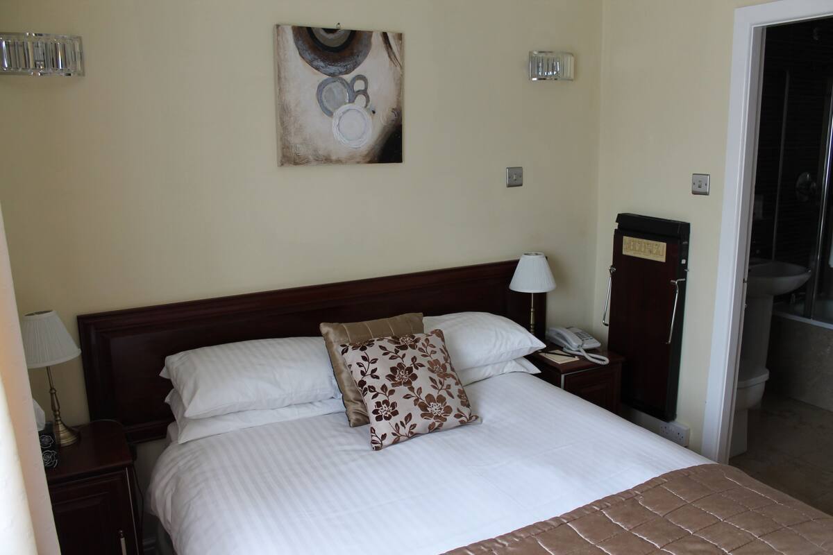 A neatly made bed with white bedding and a floral pillow, a bedside lamp, a telephone, and a wall-mounted artwork.