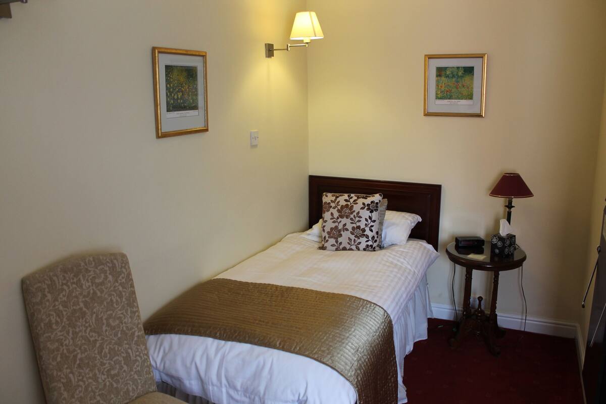 A hotel room with a bed, a chair, a bedside table with a lamp, and two framed pictures on the wall.