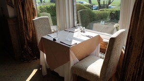 A dining table set with a white tablecloth, two chairs, and a window with a view of a parking lot and greenery. - Longview Hotel (Knutsford)