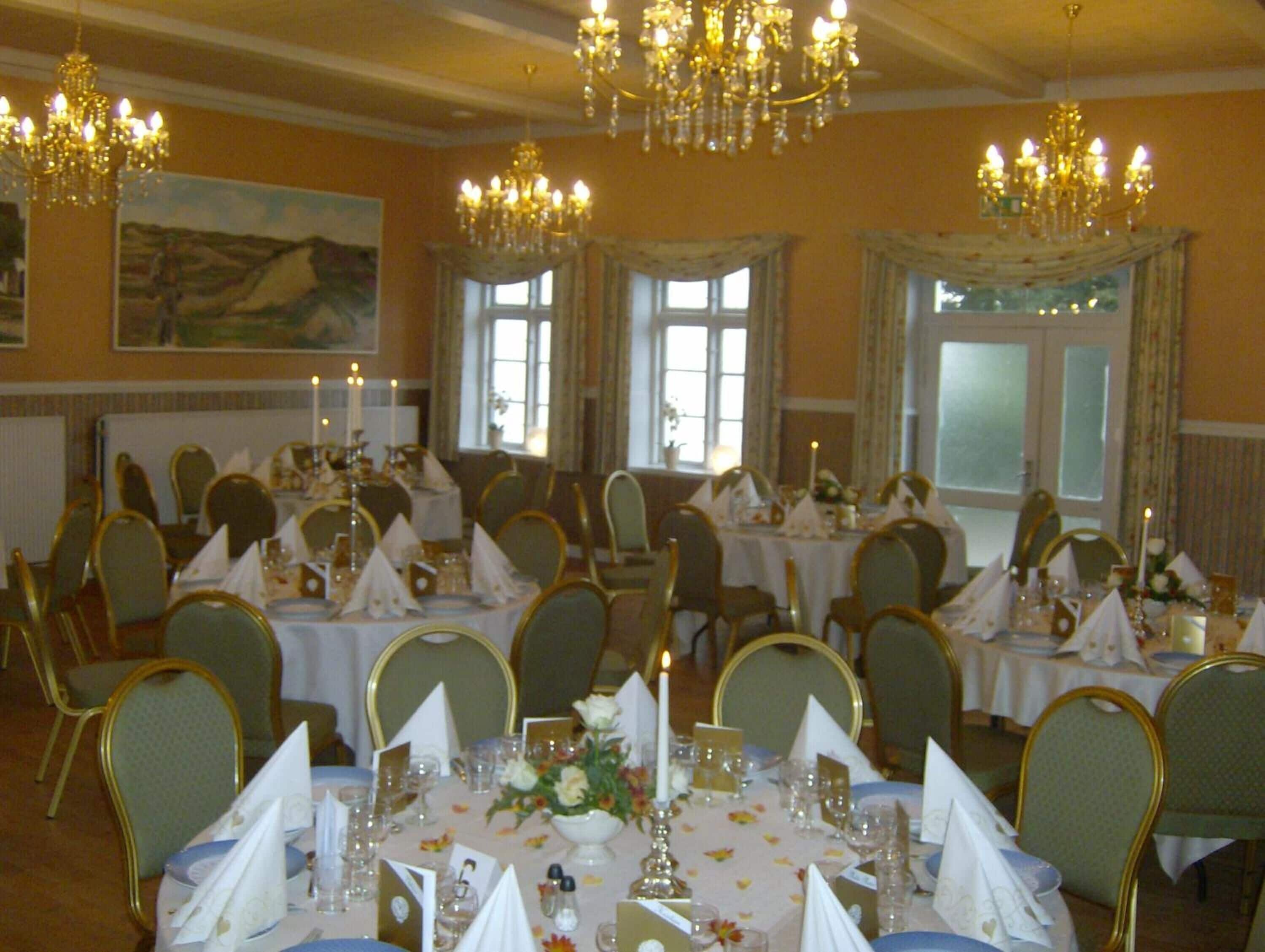 A banquet hall with round tables, chairs, chandeliers, and a large painting on the wall.
