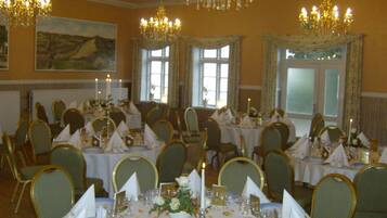 A banquet hall with round tables, chairs, chandeliers, and a large painting on the wall.