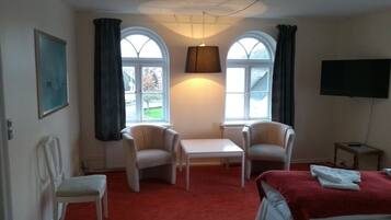 Ein Hotelzimmer mit zwei Sesseln, einem kleinen Tisch, einem Bett mit roter Decke und zwei Fenstern mit Vorhängen.