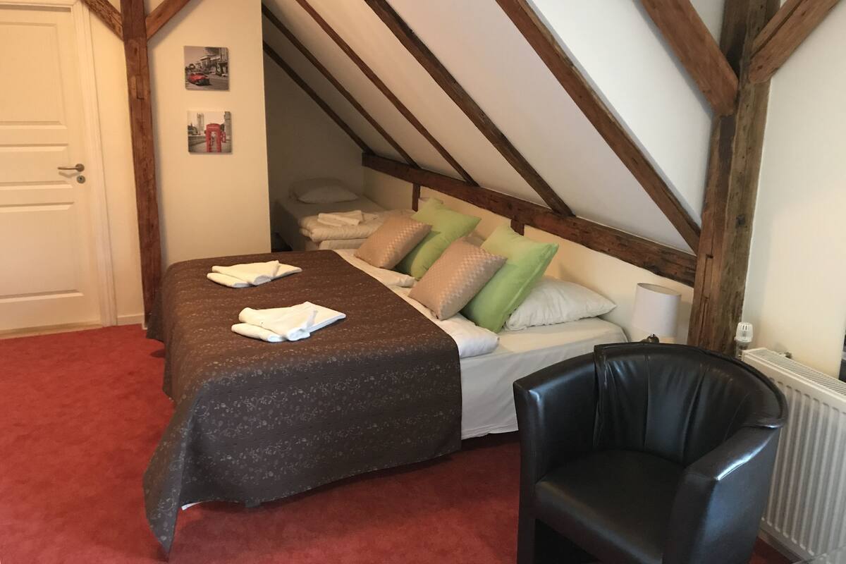 A bedroom with a sloped ceiling, a bed with multiple pillows, a black leather chair, and a radiator.