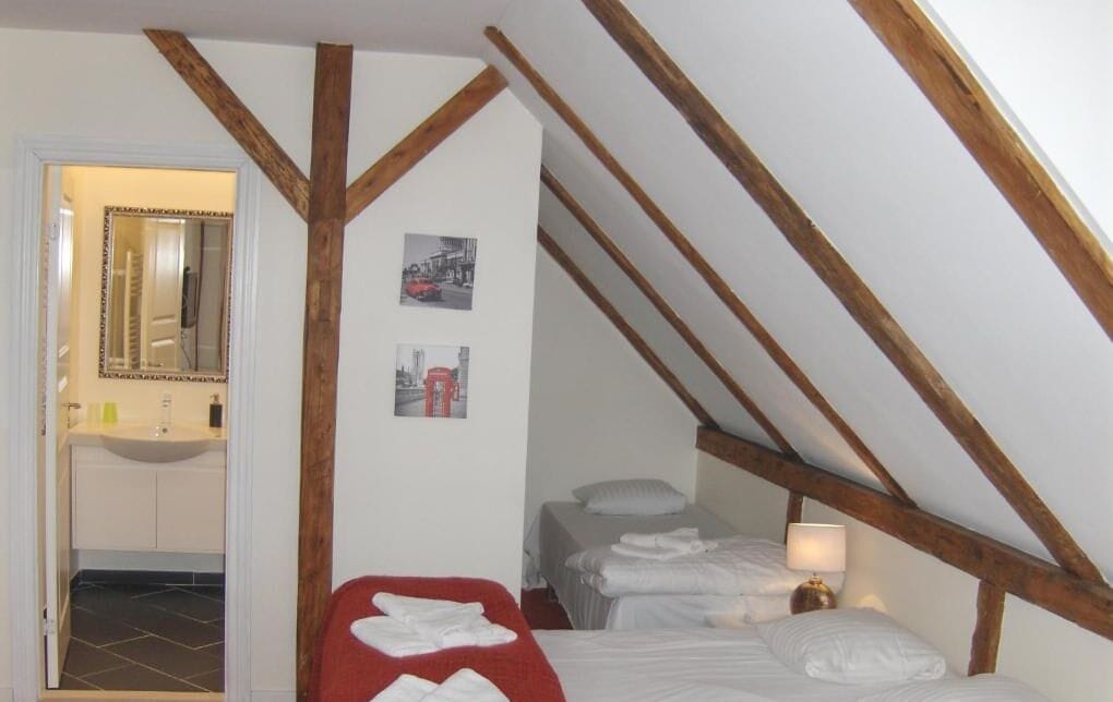 Une chambre avec un plafond en pente, un lit recouvert de draps blancs, une lampe de chevet et une petite table de chevet.