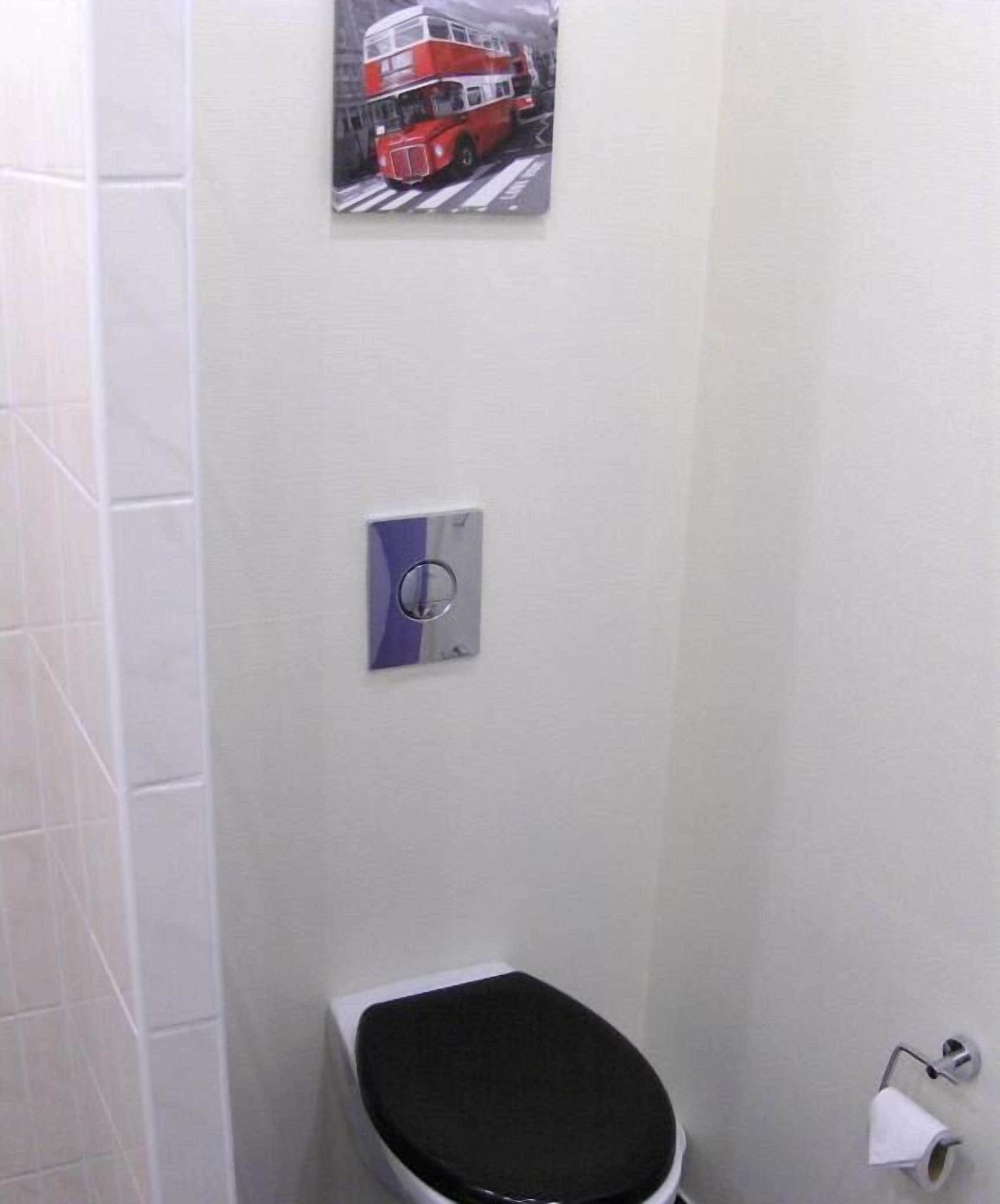 Une salle de bain avec un mur recouvert de carreaux blancs, des toilettes avec un abattant noir, un sèche-mains et une image d’un autobus à deux étages rouge.