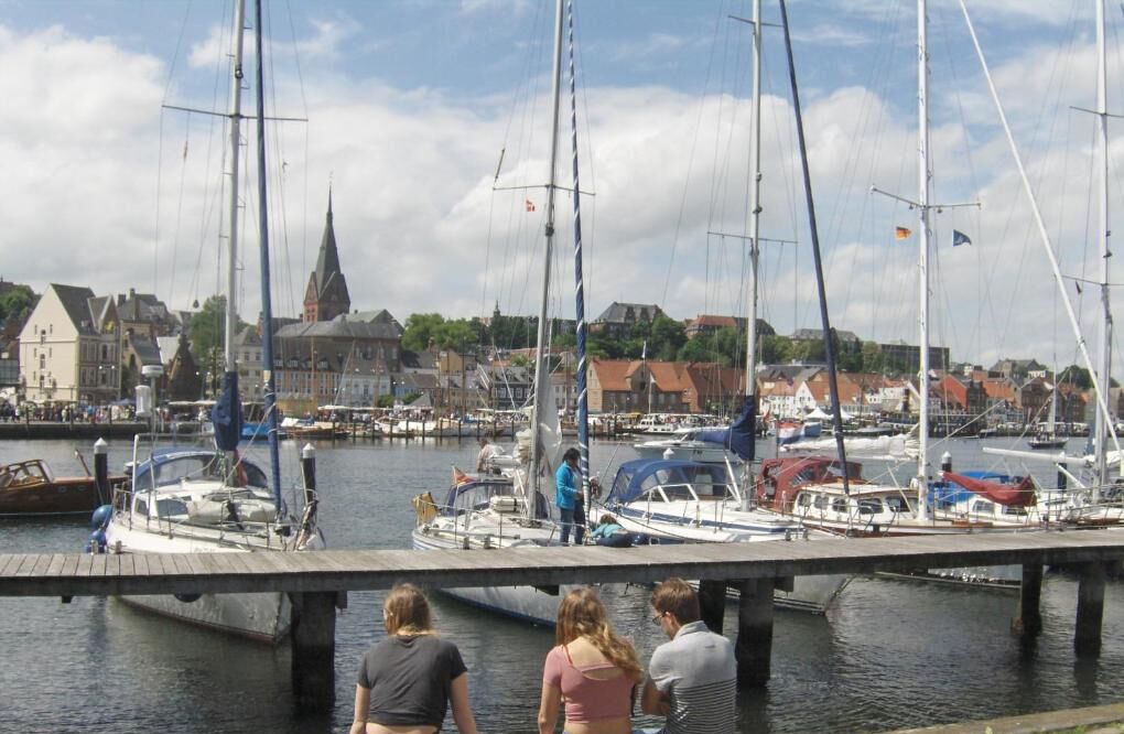 Un port de plaisance avec des bateaux amarrés à des quais en bois, une ville avec des bâtiments en arrière-plan et un ciel dégagé.