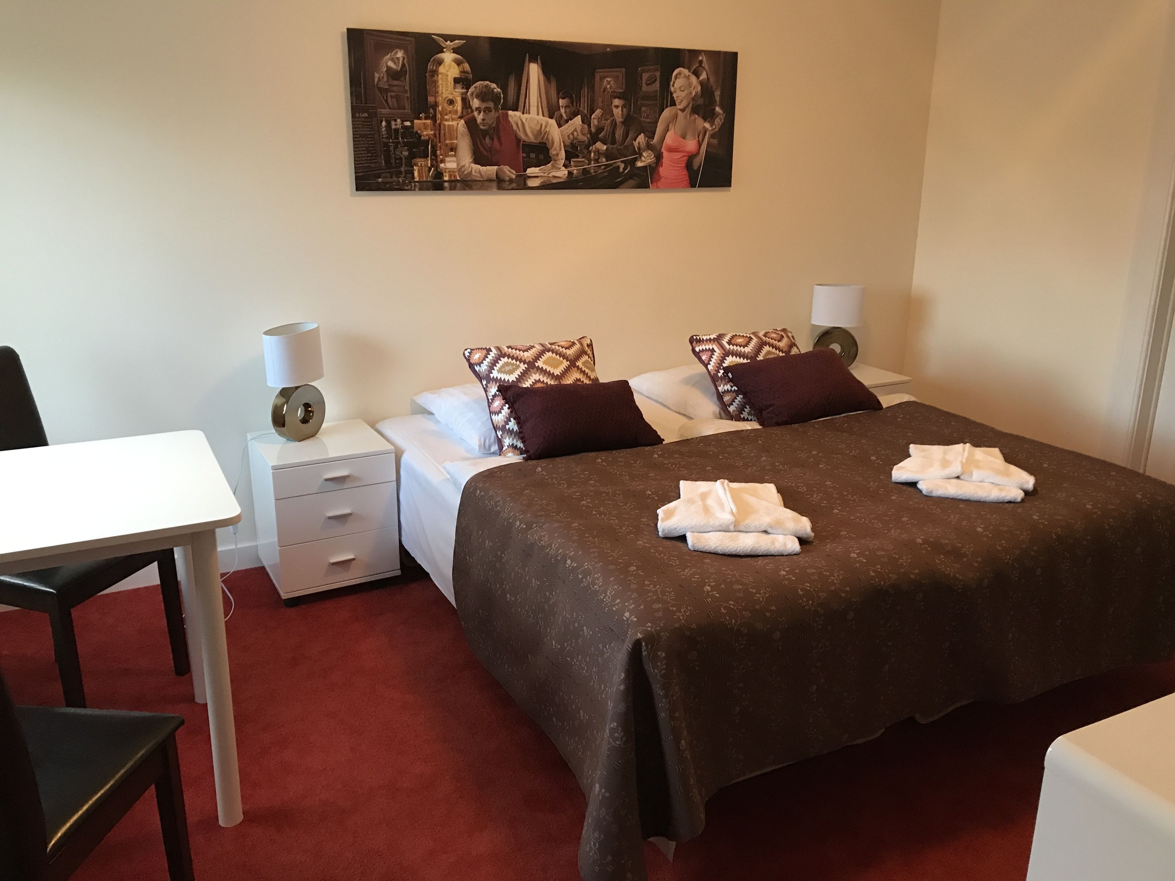 A hotel room with a bed, two bedside tables, a desk, and a chair. There is a painting on the wall above the bed.