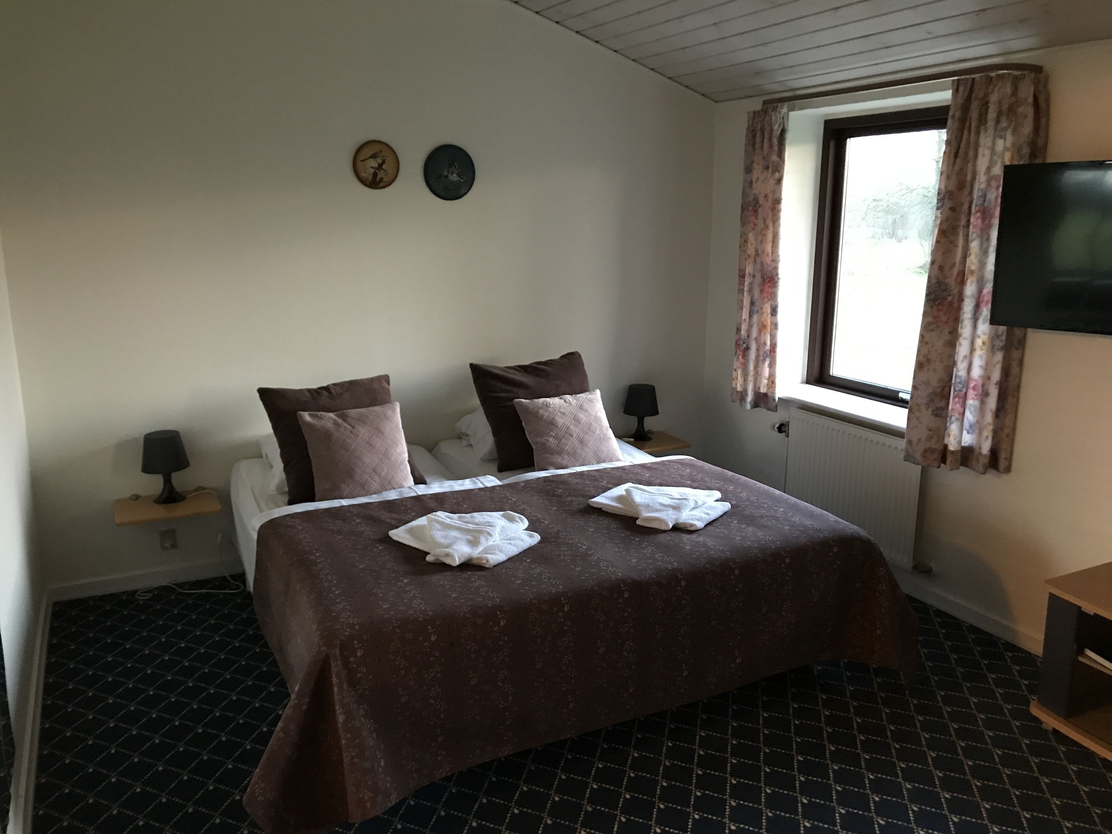 Une chambre d’hôtel avec un lit, deux oreillers, une table de chevet avec une lampe, un téléviseur et une fenêtre avec des rideaux.