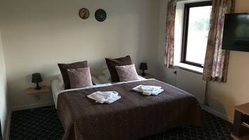 Une chambre d’hôtel avec un lit, deux oreillers, une table de chevet avec une lampe, un téléviseur et une fenêtre avec des rideaux.